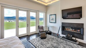 A modern bedroom with a large bed, a fireplace, and a view of a green landscape through large windows. - Motorcycle-themed Home in Spearfish w/ Hot Tub! (Spearfish)