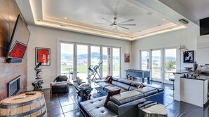 A spacious living area with a large sectional sofa, a coffee table, and a TV mounted on the wall. There is a wooden barrel, a ceiling fan, and large windows offering a view of the outdoors. - Motorcycle-themed Home in Spearfish w/ Hot Tub! (Spearfish)