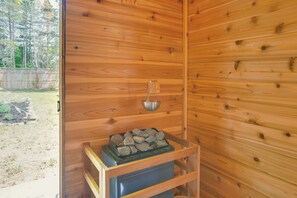 Ein Holzblockhaus-Innenraum mit Kamin, einem Metallbehälter auf dem Sims und Blick ins Freie.