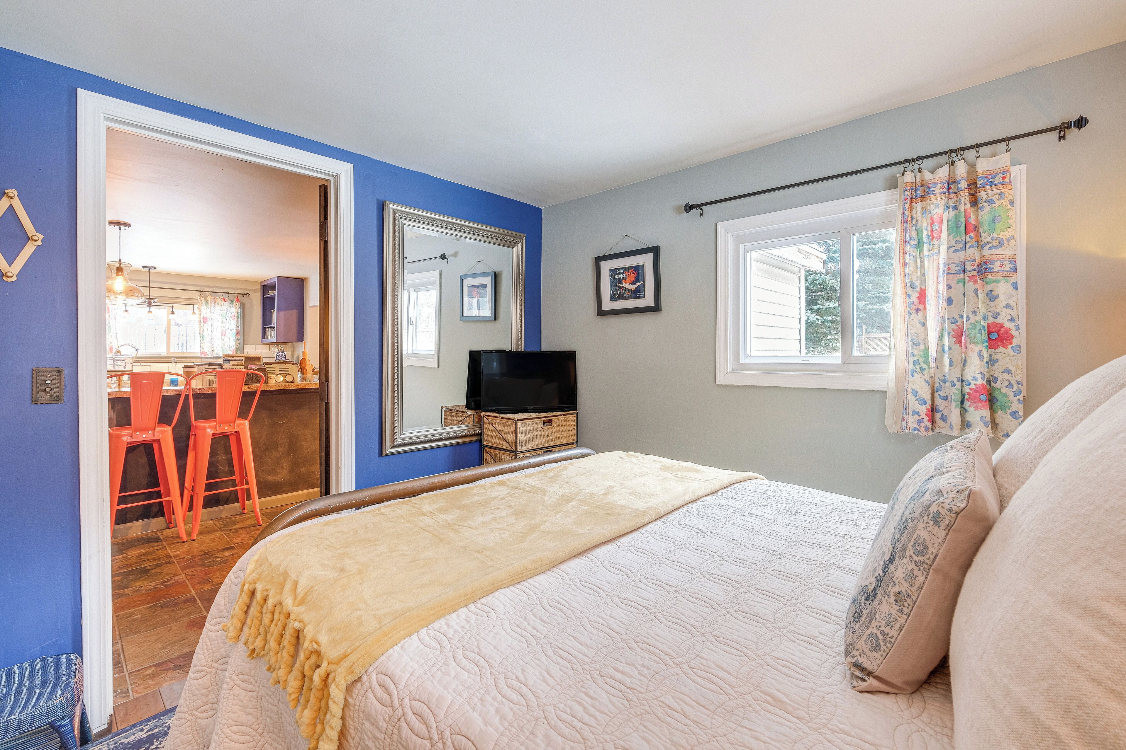 A bedroom with a bed, a TV, a window with curtains, and a view of a kitchen and dining area.