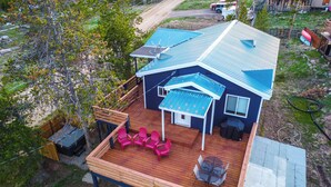 Una casa con tetto blu, un patio con pavimento in legno e una zona porticata con sedie rosa e un tavolo.