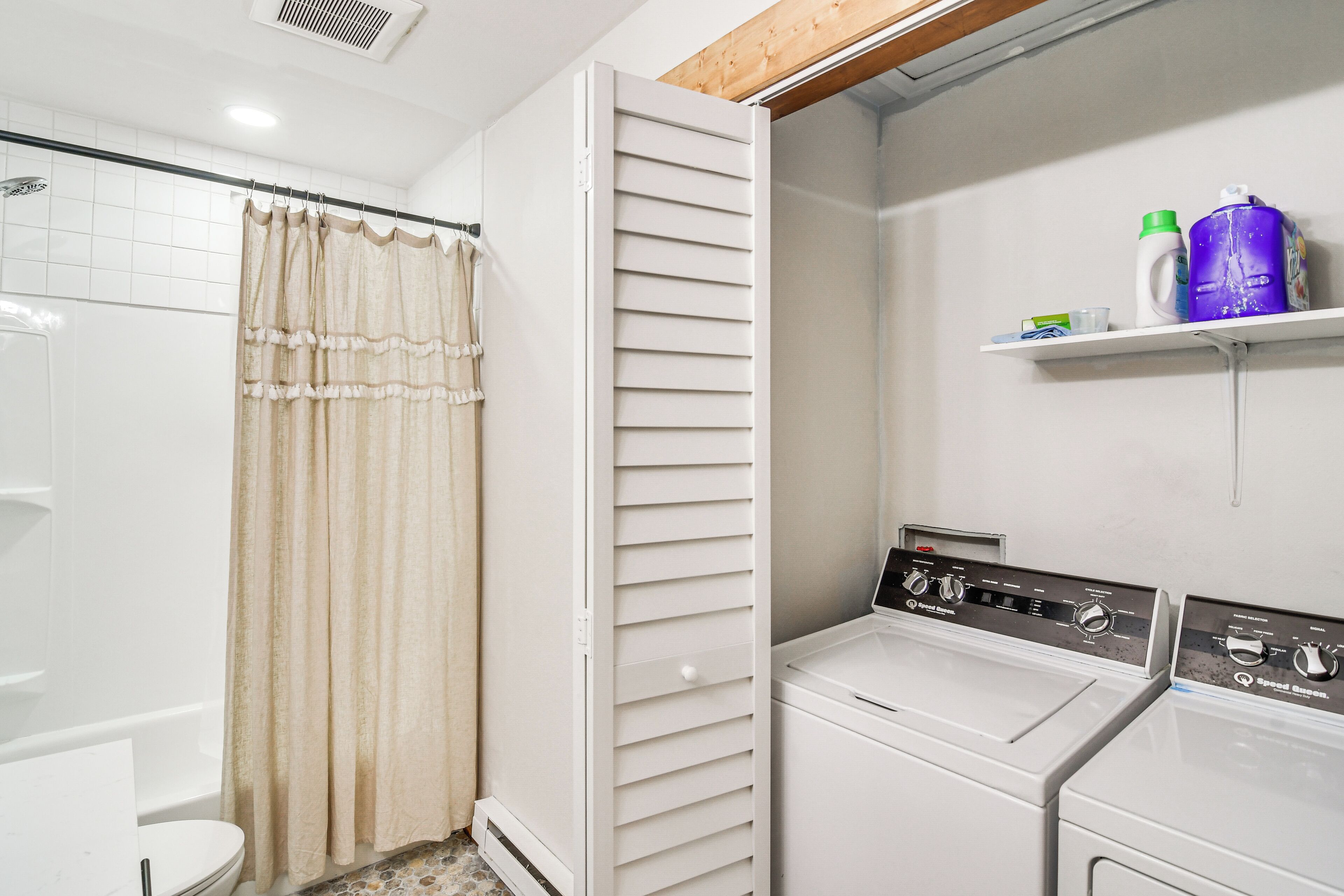 A laundry area with two white washing machines, a shower with beige curtains, and a shelf with cleaning supplies.