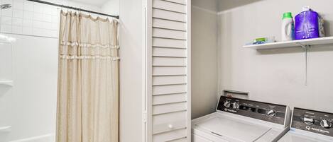 A laundry area with two white washing machines, a shower with beige curtains, and a shelf with cleaning supplies.