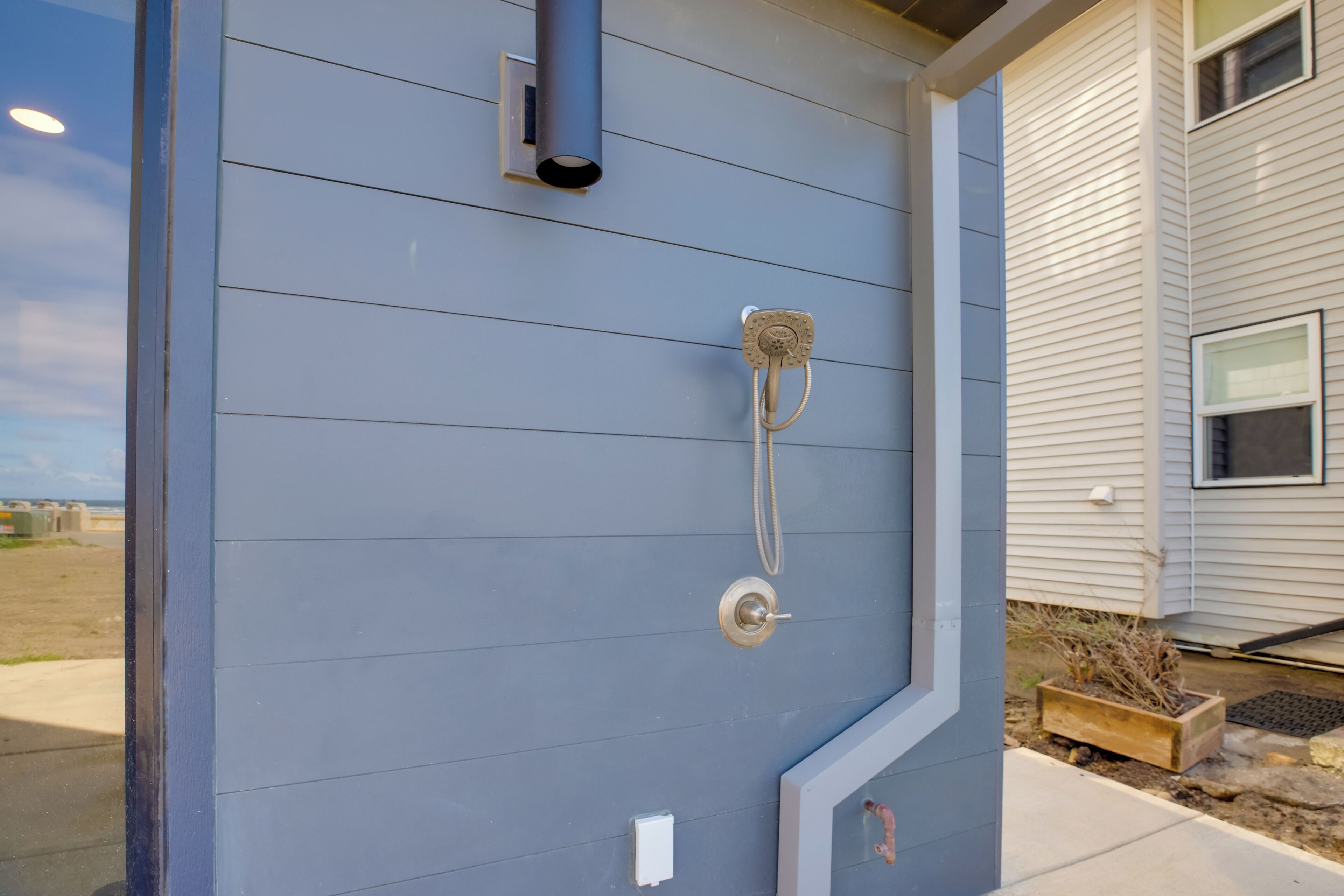 Pared exterior gris con un cabezal de ducha metálico y un pequeño cuadro eléctrico blanco.