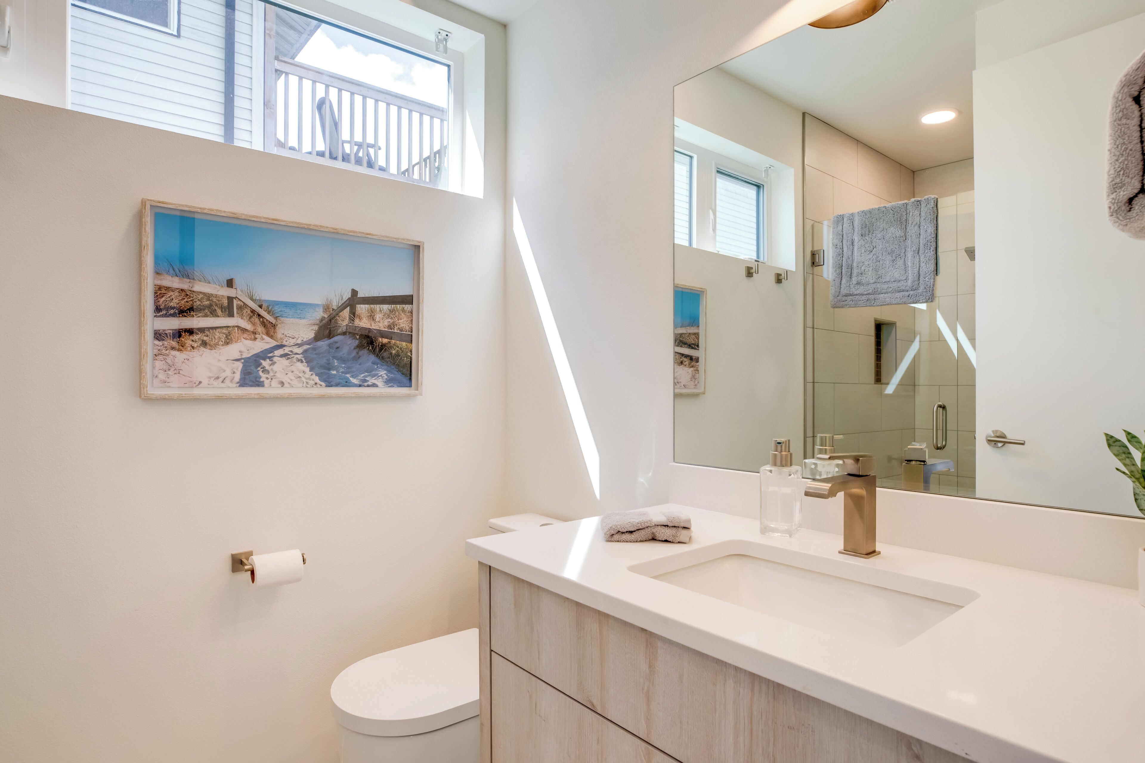 Une salle de bain moderne avec un grand miroir, un lavabo blanc et des toilettes. Un tableau représentant une scène de plage est accroché au mur.