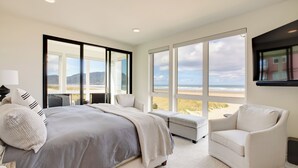 Un dormitorio con una cama grande, un sofá y un televisor. Amplias ventanas ofrecen vistas a la playa y a las montañas.