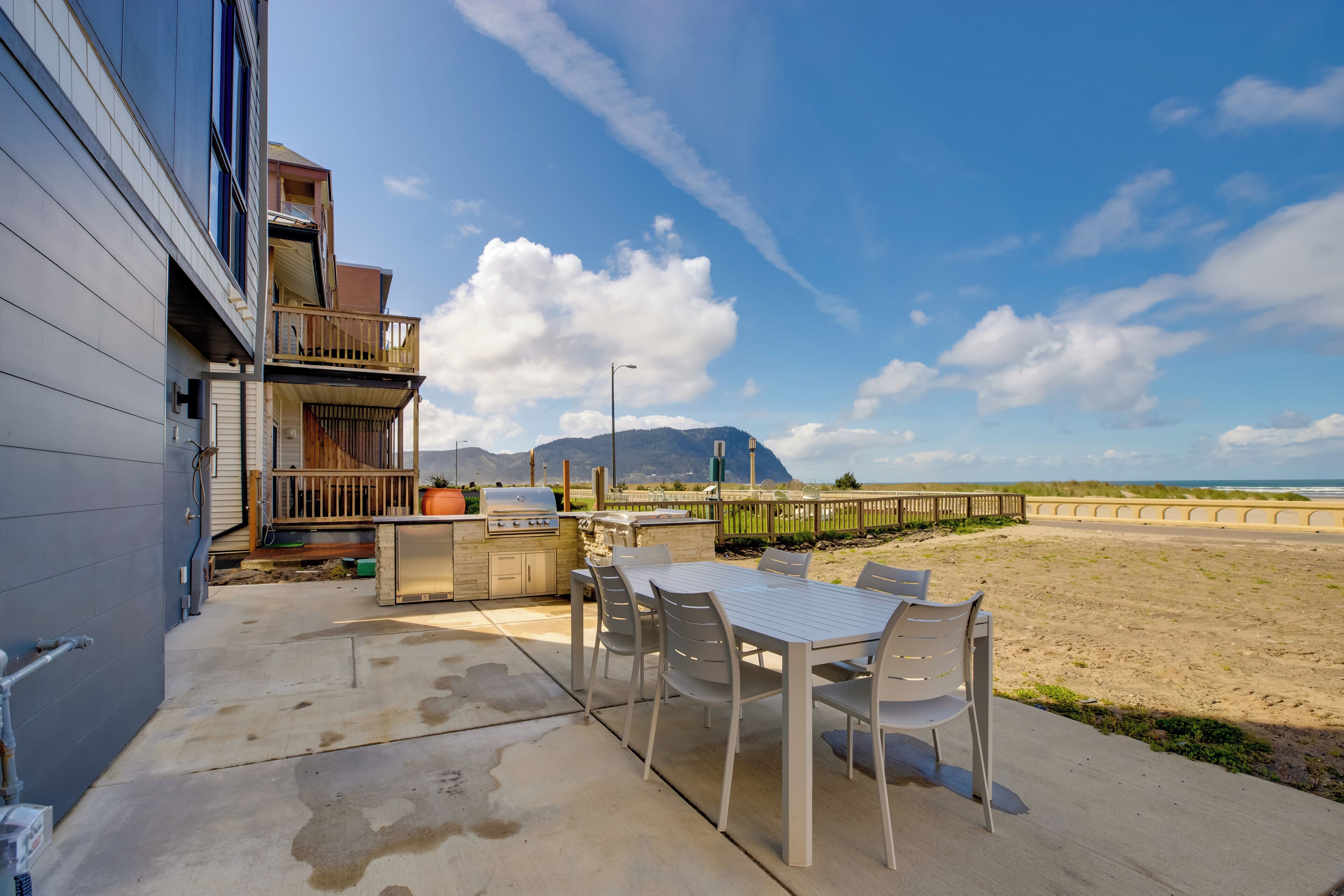 A patio with a table and chairs, a barbecue grill, and a view of the beach and mountains.