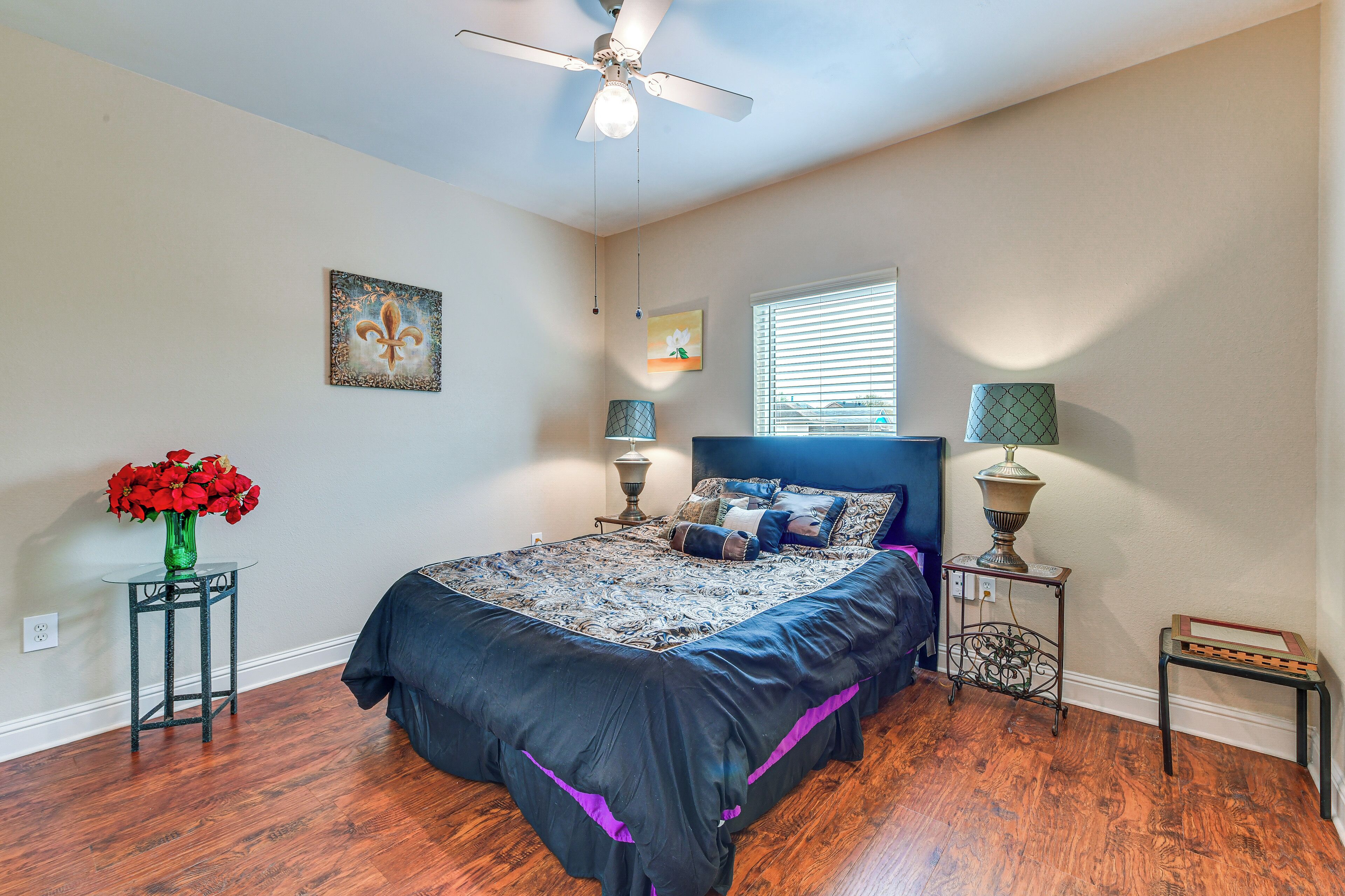 A bedroom with a bed, a ceiling fan, a window with blinds, a vase with flowers, and a lamp on a side table.