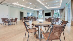 A spacious conference room with round tables and wicker chairs, large windows with curtains, and a flat-screen TV on the wall.