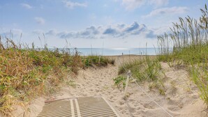 A sandy beach path leading to the sea, with grass and a drainage grate.