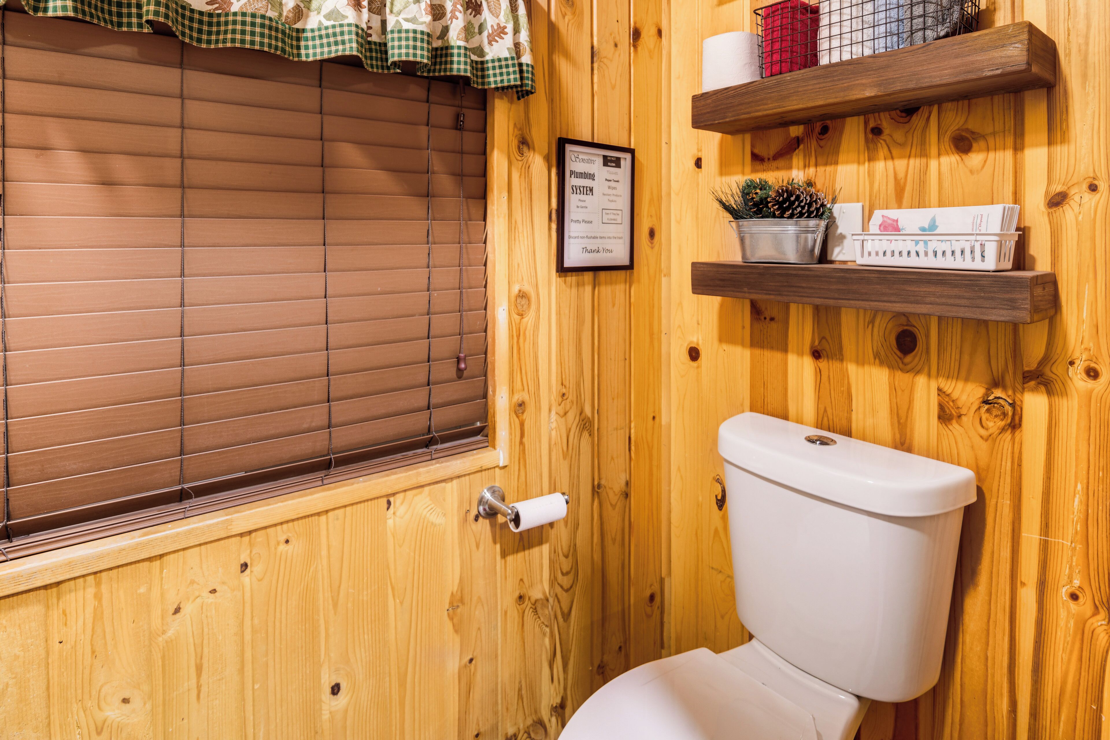 Et badeværelse i træ med toilet, et vindue med persienner og hylder med toiletartikler.
