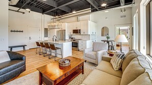 Un amplio salón con una mesa de centro de madera, un sofá y un sillón a rayas. Se ve una cocina al fondo, con armarios blancos y electrodomésticos de acero inoxidable.