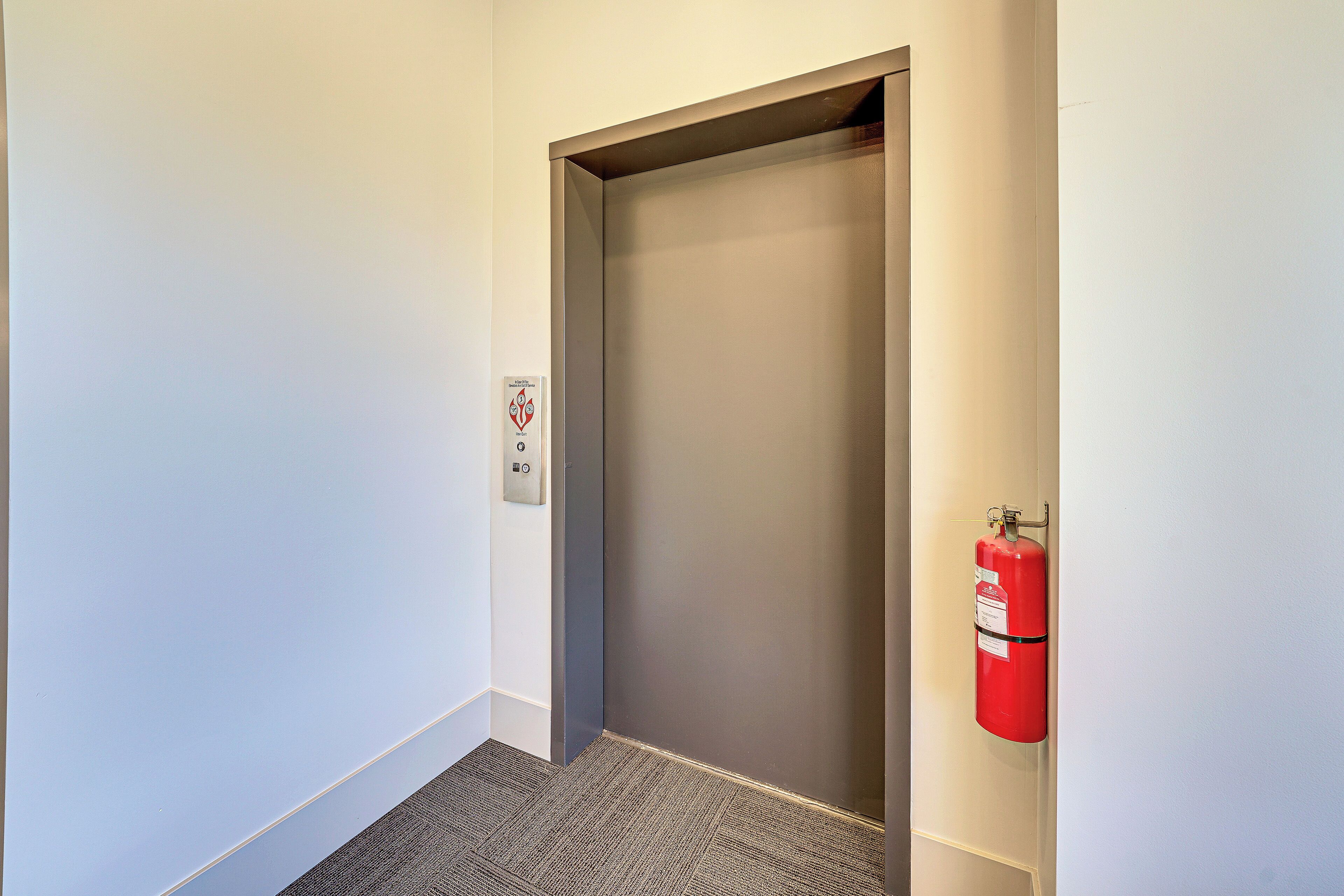 Un couloir étroit avec une porte deascenseur fermée, un extincteur fixé au mur et un sol recouvert de moquette.