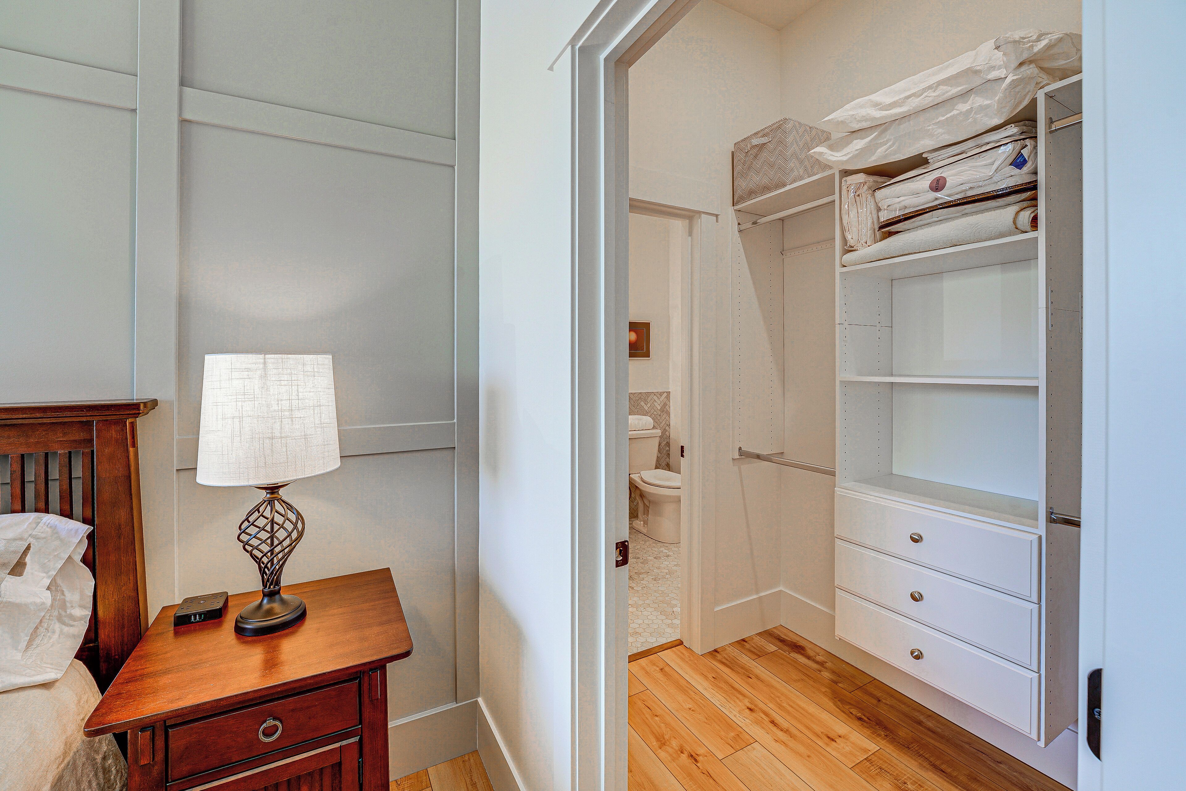 A bedroom with a wooden nightstand, a lamp, a bed, and a closet with shelves and drawers.