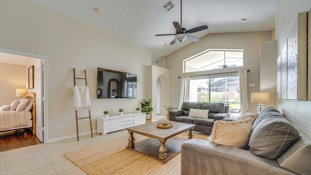A spacious living room with a large window, a ceiling fan, and a flat-screen TV mounted on the wall.