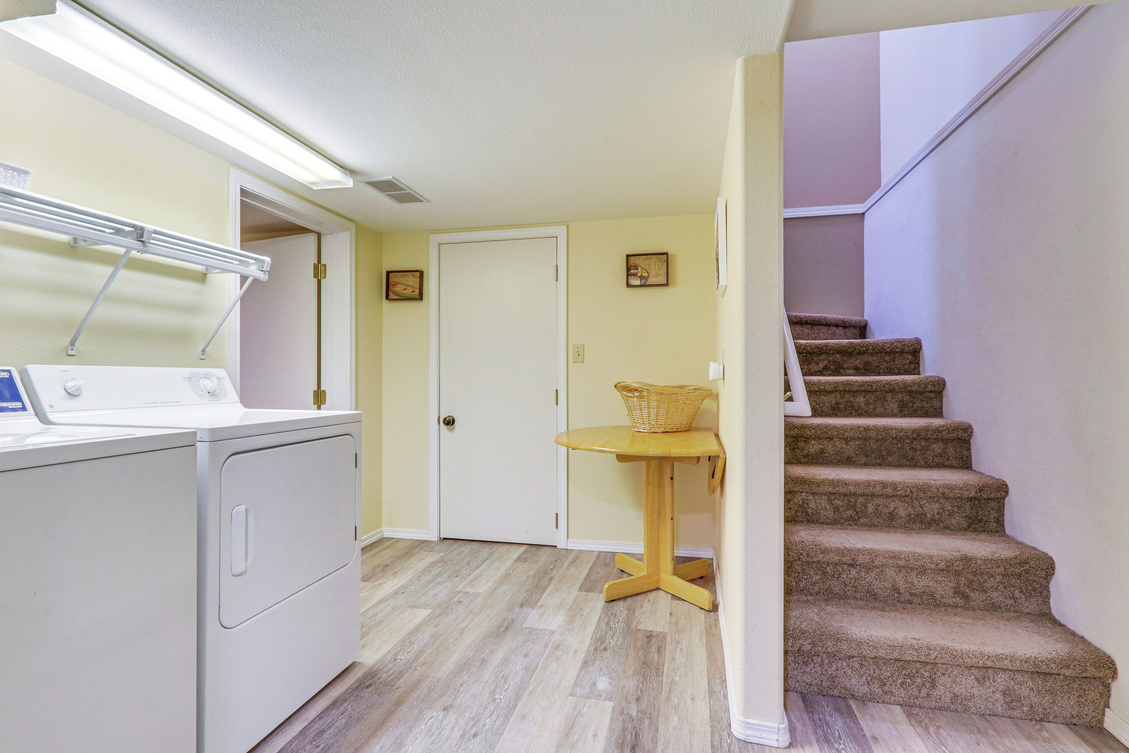 A laundry area with a washing machine and dryer, a small table with a basket, and a carpeted staircase.