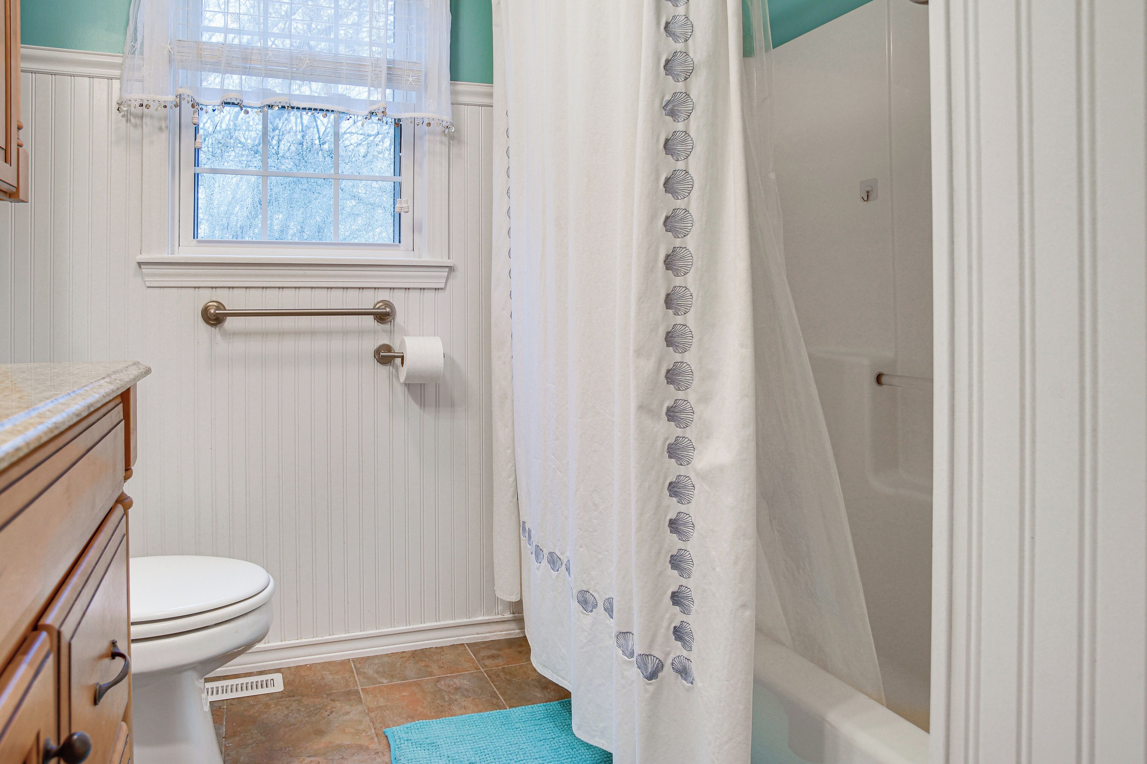 Une salle de bain avec des toilettes, une fenêtre avec un rideau, un rideau de douche orné de motifs de coquillages et un tapis de bain bleu.