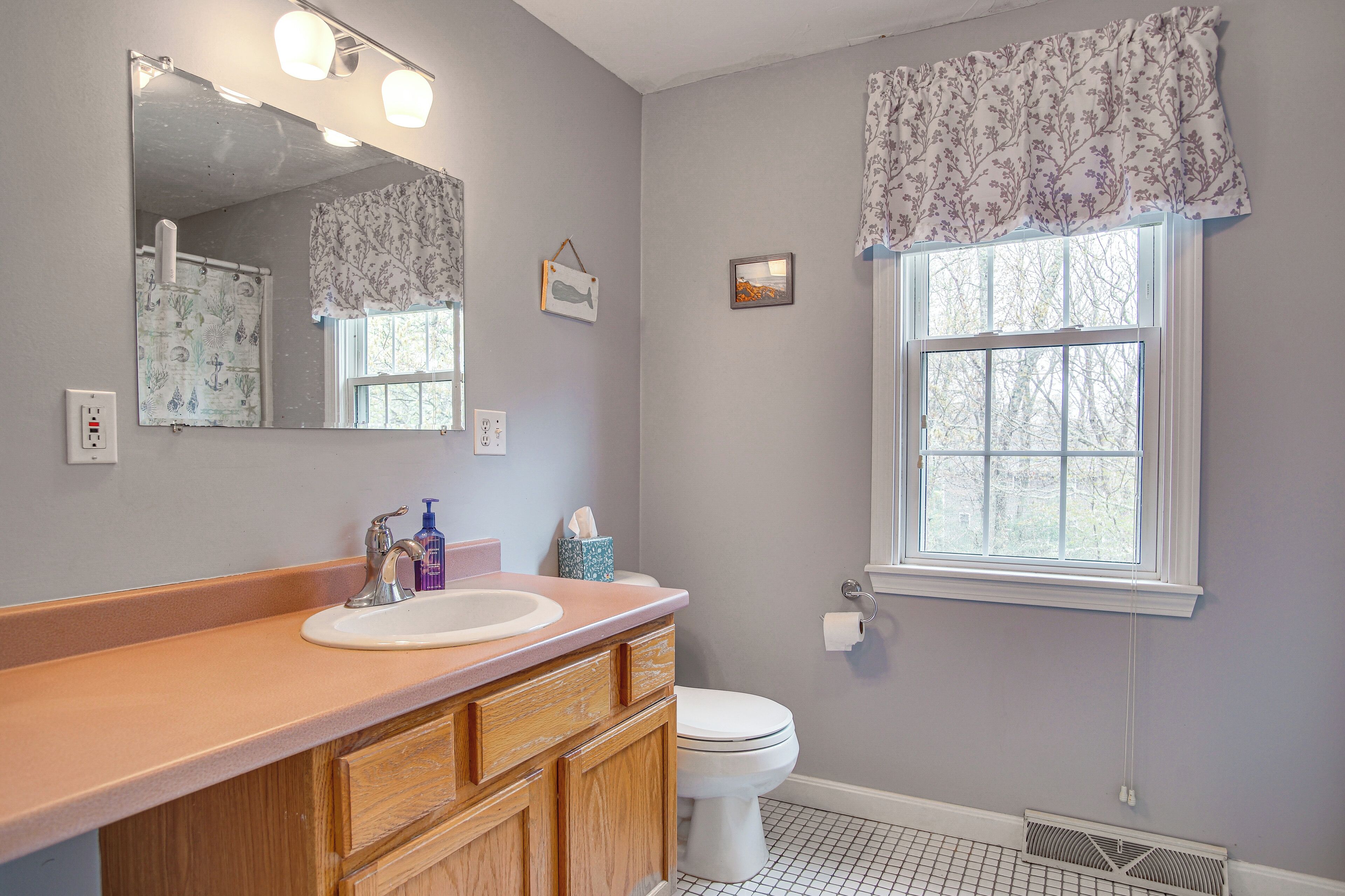 A bathroom with a wooden vanity, a white sink, a mirror, a toilet, and a window with a floral valance.