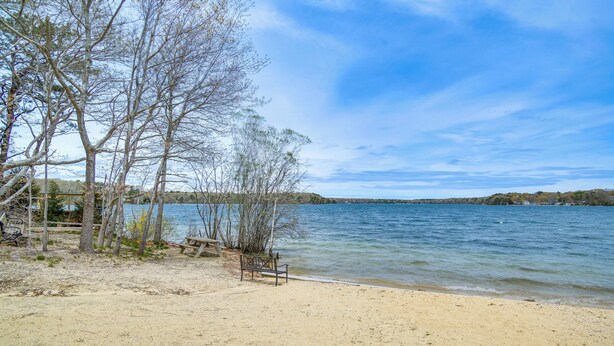 A sandy beach with a wooden bench and trees, overlooking a calm lake. - Beach & Lake Access: Group Getaway in Mashpee! (Mashpee)
