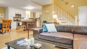 A spacious living area with a leather sofa, wooden dining table, and a kitchen in the background. - Beach & Lake Access: Group Getaway in Mashpee! (Mashpee)
