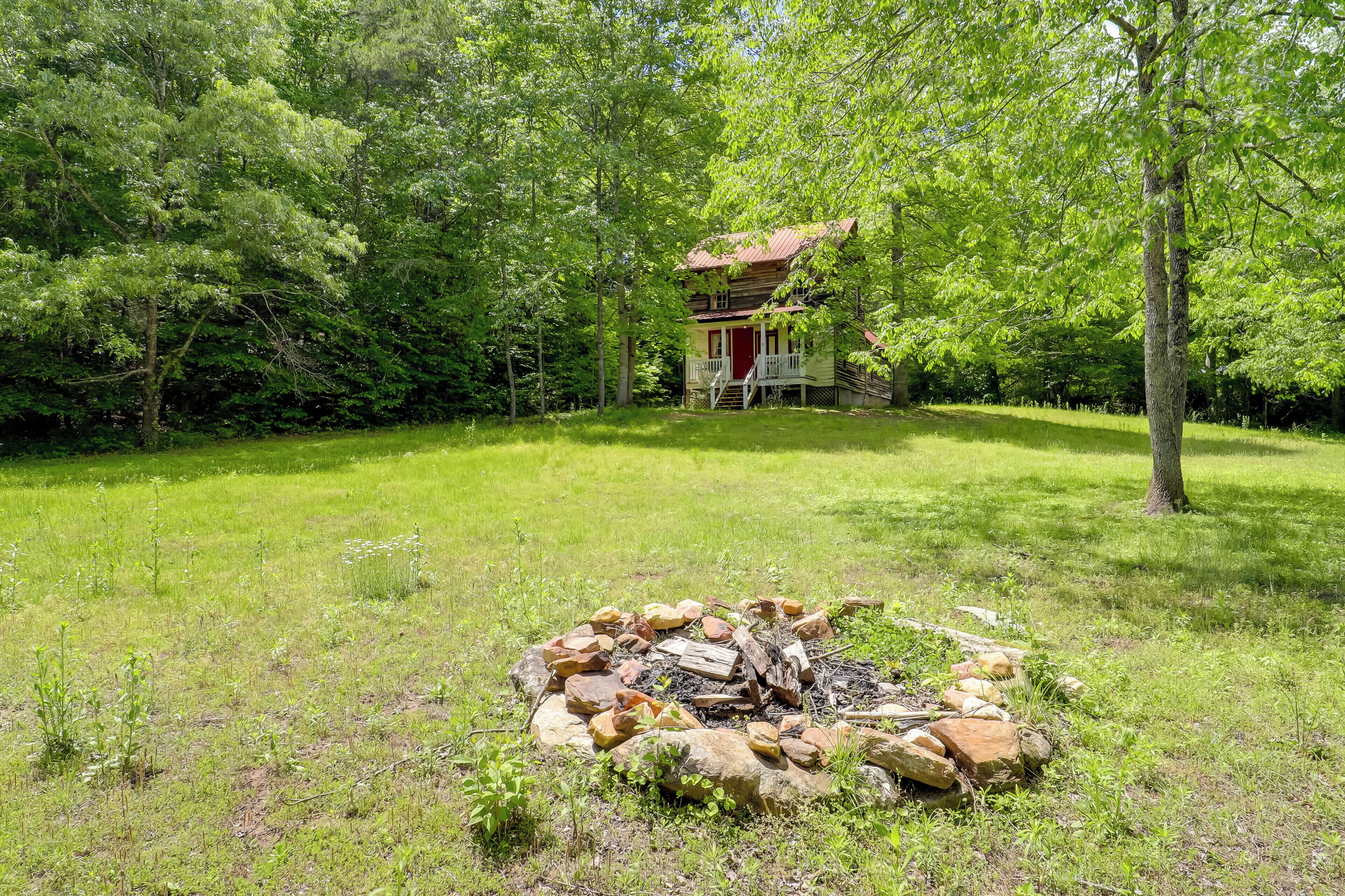 A cabin surrounded by dense forest, a fire pit in the foreground, and a red door.
