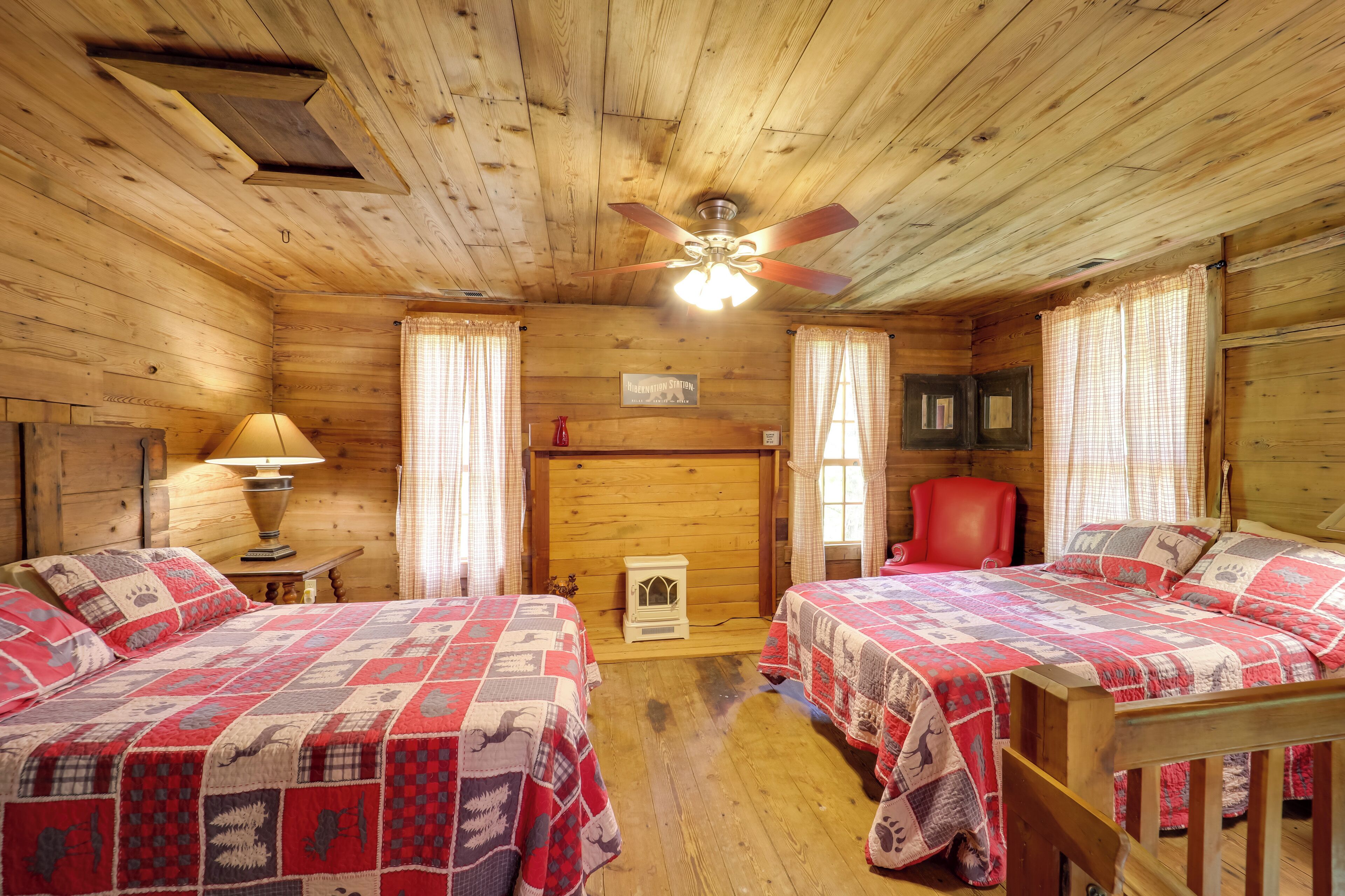 A cabin-style room with two beds, wooden walls, a ceiling fan, a fireplace, and a red chair.