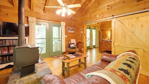 A cozy cabin interior with a stone fireplace, leather sofa, wooden walls, and a television. - Hot Tub & Trail Access: 92-acre Retreat in Marion! (Marion)
