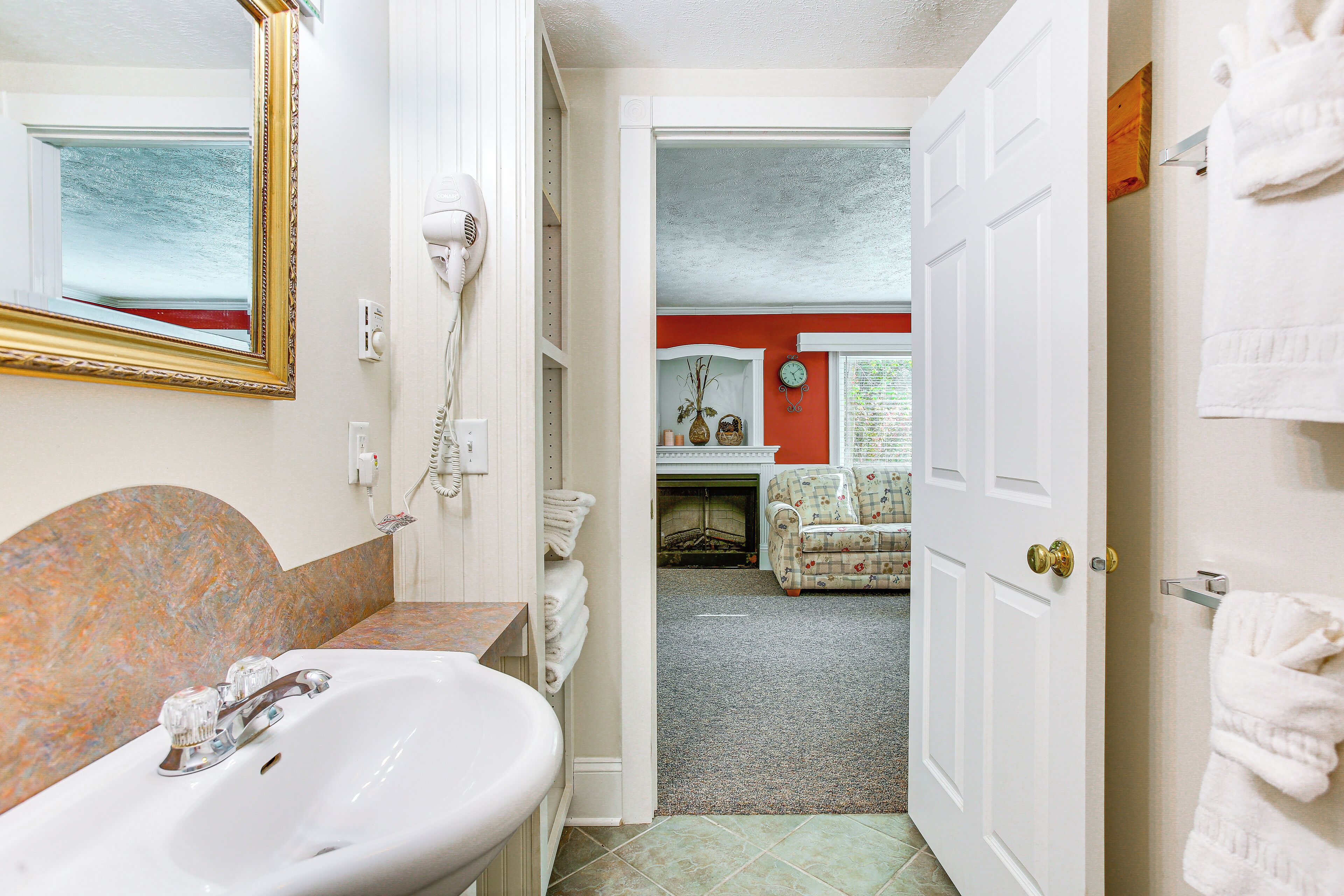 A bathroom with a sink, mirror, and hairdryer. There is a door leading to a living area with a couch and a clock on the wall.