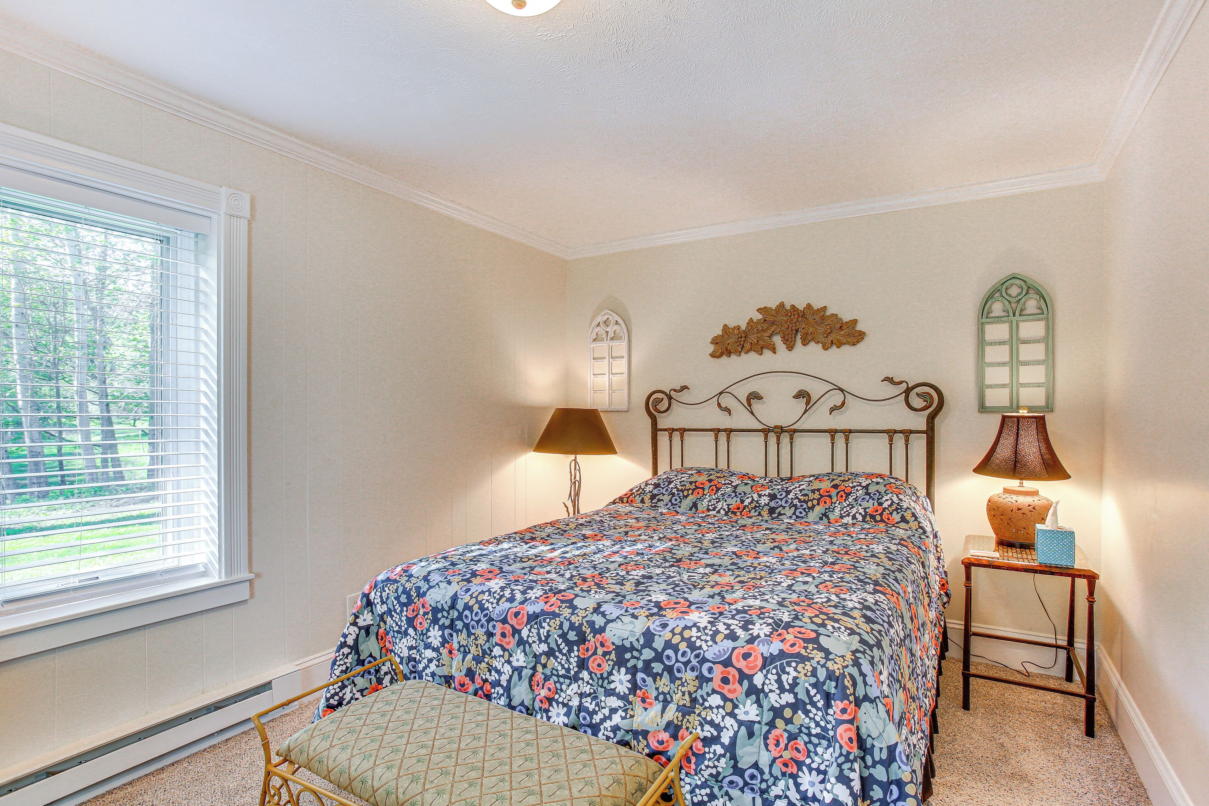 A bedroom with a floral bedspread, a wooden headboard, a nightstand with a lamp, and a window with blinds.