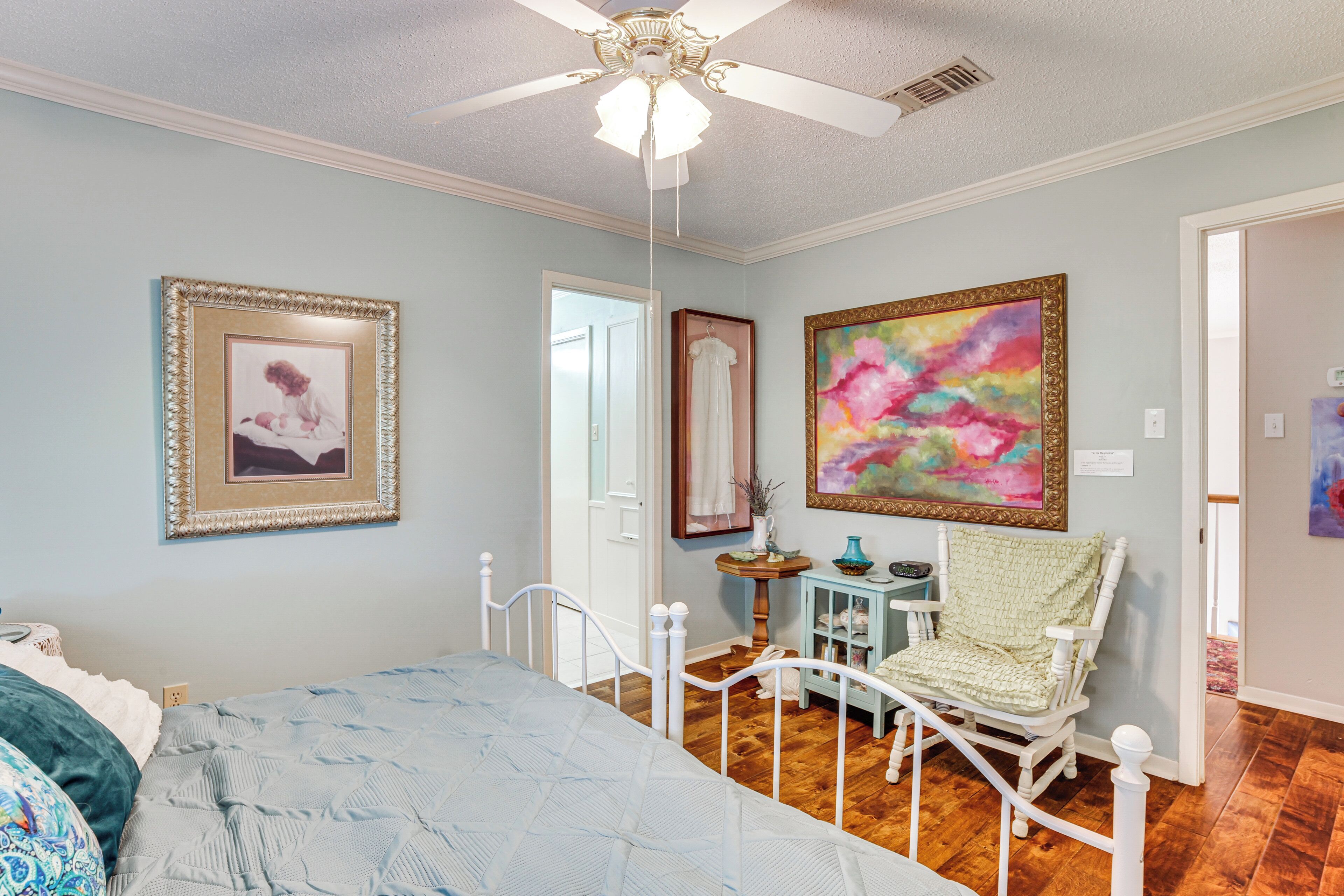 A bedroom with a bed, a chair, a ceiling fan, and a painting on the wall.