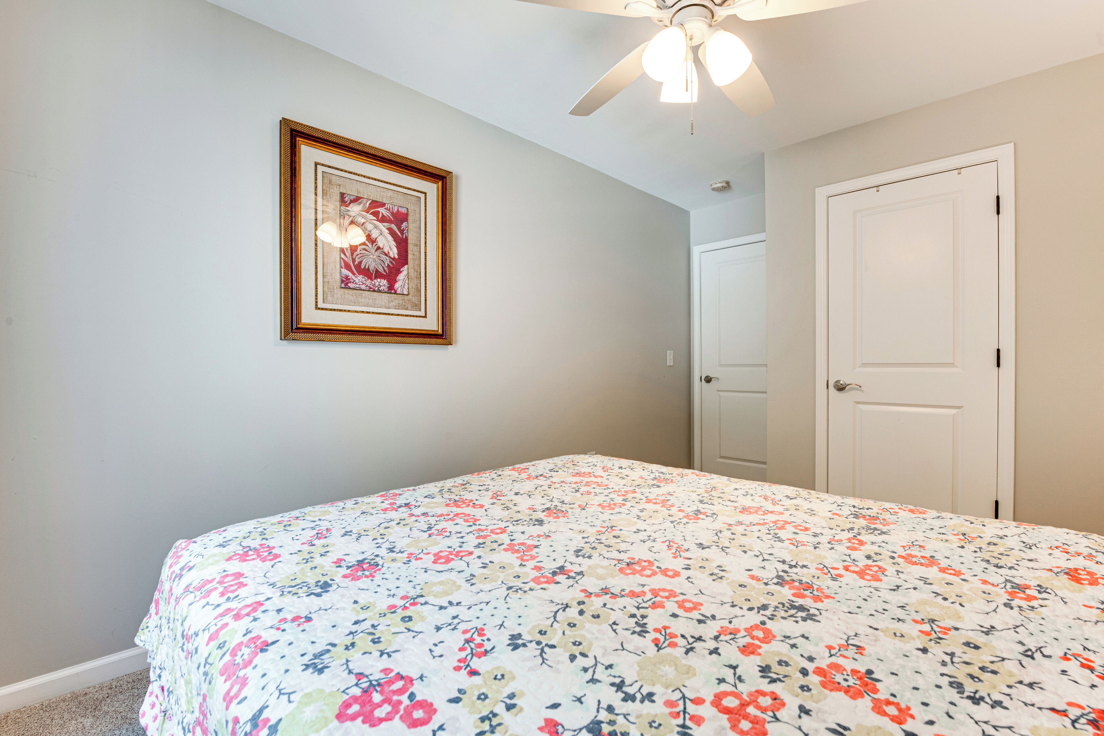 Un dormitorio con una colcha estampada con motivos florales, un ventilador de techo, un cuadro enmarcado en la pared y una puerta cerrada.