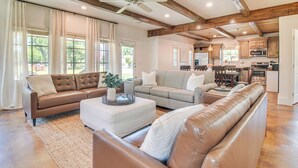 A spacious living room with a brown leather sofa, a grey sofa, and a white ottoman. There is a ceiling fan, wooden beams, and a kitchen in the background.