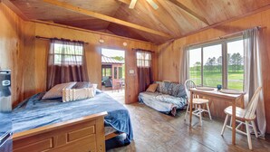 A wooden cabin with a bed, a sofa, a dining table with chairs, and large windows offering a view of the outdoors. - Dog-friendly Cabin Near Pymatuning Lake! (Kinsman)