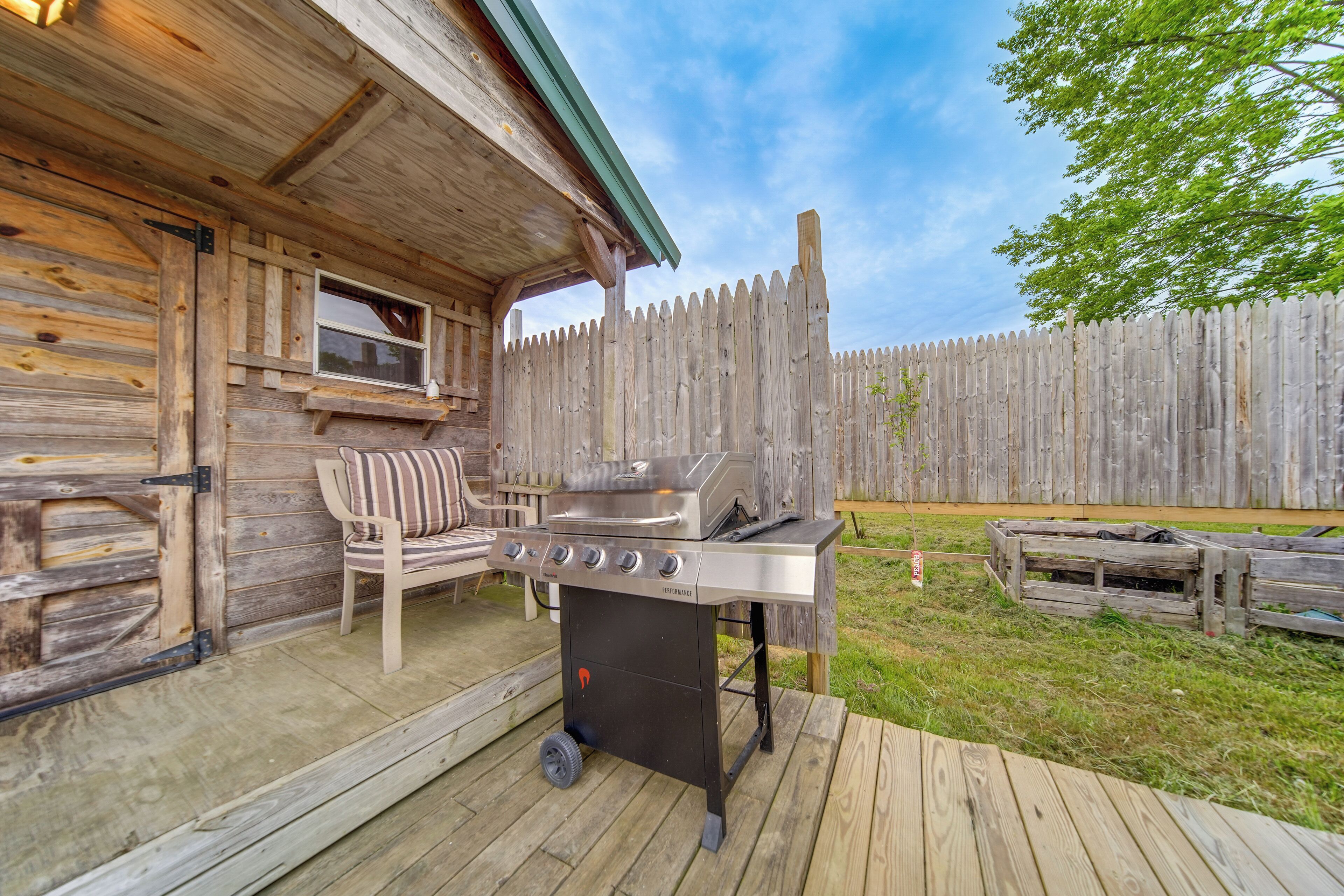 Una terraza de madera con una parrilla, un área de estar exterior con cojines y una cerca de madera al fondo.