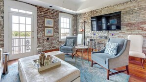 A living room with a brick wall, a flat-screen TV, and two armchairs. - The Riverfront Loft, Charming | River Views (Savannah)