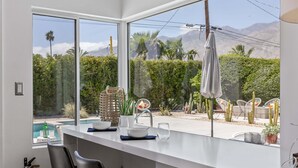 A modern kitchen with large windows, a pool view, and outdoor seating. - Desert Bloom Retreat - Upscale Mid-Mod Home! (Palm Springs)