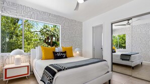 A modern bedroom with a large bed, a nightstand, a mirror, and a view of greenery outside. - Desert Bloom Retreat - Upscale Mid-Mod Home! (Palm Springs)