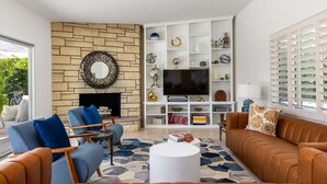 A living room with a fireplace, a television, and a bookshelf. - Desert Bloom Retreat - Upscale Mid-Mod Home! (Palm Springs)