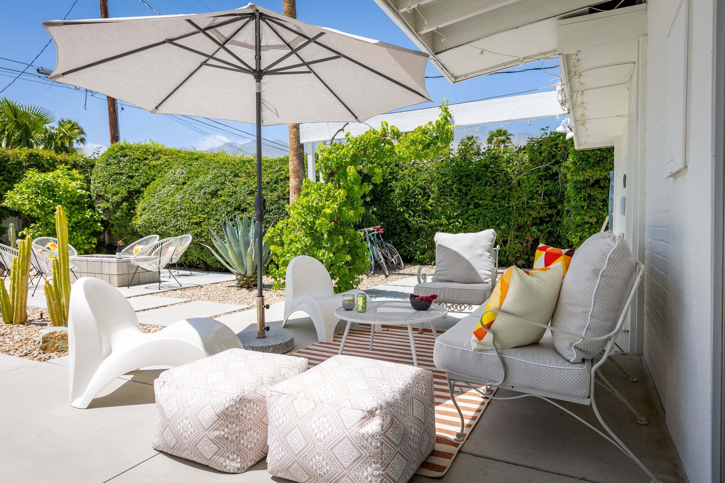 Un patio exterior con mobiliario blanco, incluyendo un sofá y sillas, una mesa con un cuenco y una bebida, y una sombrilla grande que proporciona sombra.