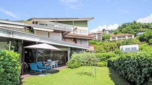 Ein modernes Haus mit überdachter Terrasse, kleinem Rasen und Blick auf benachbarte Gebäude.