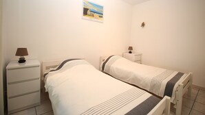 Une petite chambre propre, avec deux lits simples, une table de chevet avec une lampe, une commode et un tableau encadré au mur.