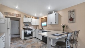 A modern kitchen with white cabinets, a central island, and a dining area with two chairs. - Serene Riverside Retreat 3 Bedroom Home by RedAwning (Little Suamico)
