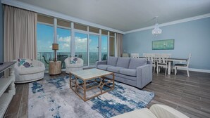 A spacious living room with a large window, a grey sofa, a wooden coffee table, and a dining area with white chairs and a table.