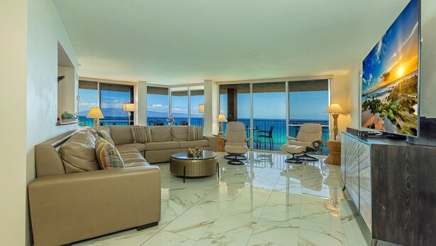A spacious living room with a large sofa, armchairs, and a coffee table. The room has large windows offering a view of the ocean. - Royal Kahana 1211 2 Bedroom Condo (Lahaina)