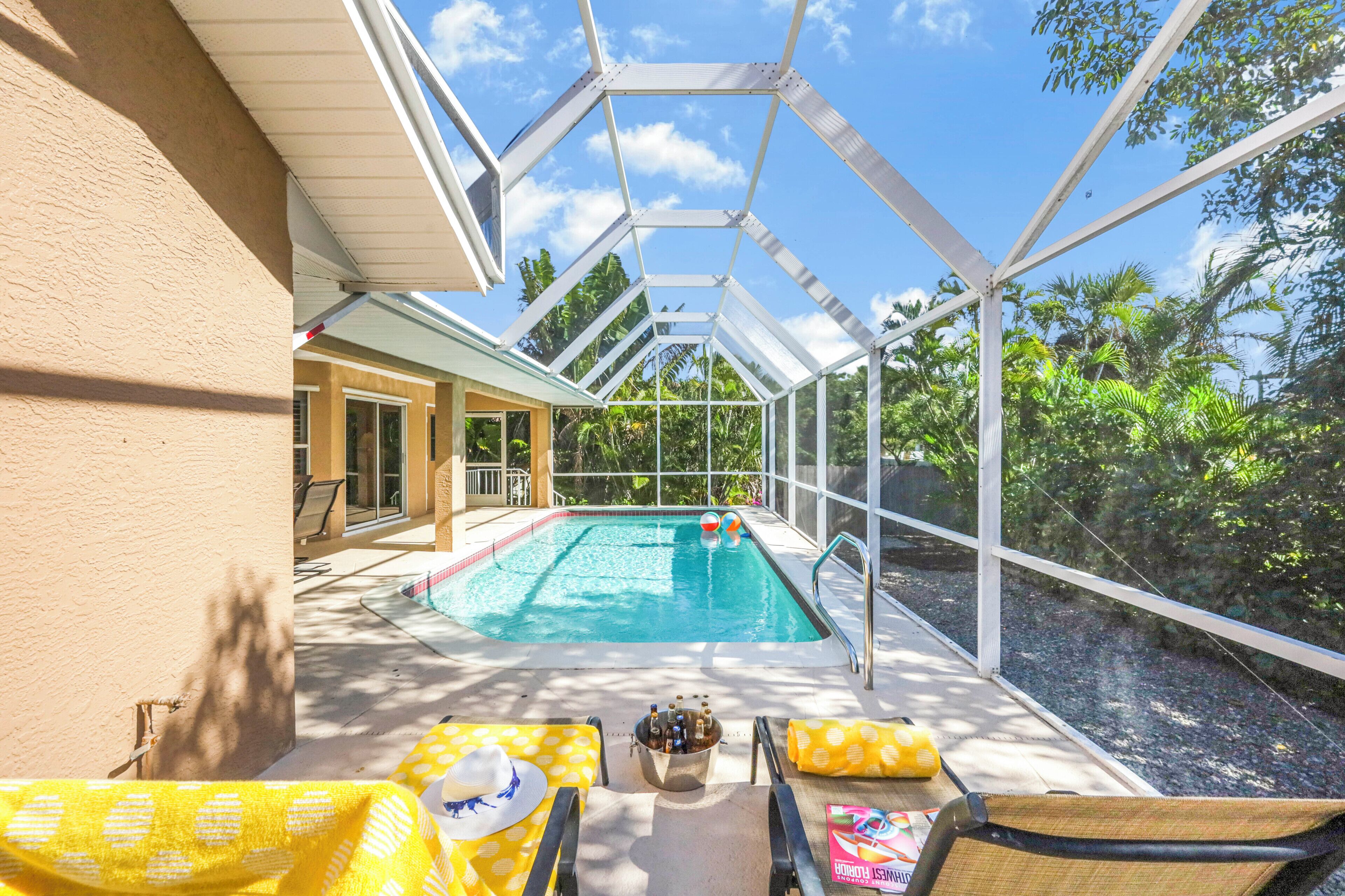 A covered outdoor swimming pool with lounge chairs and a table, surrounded by greenery.