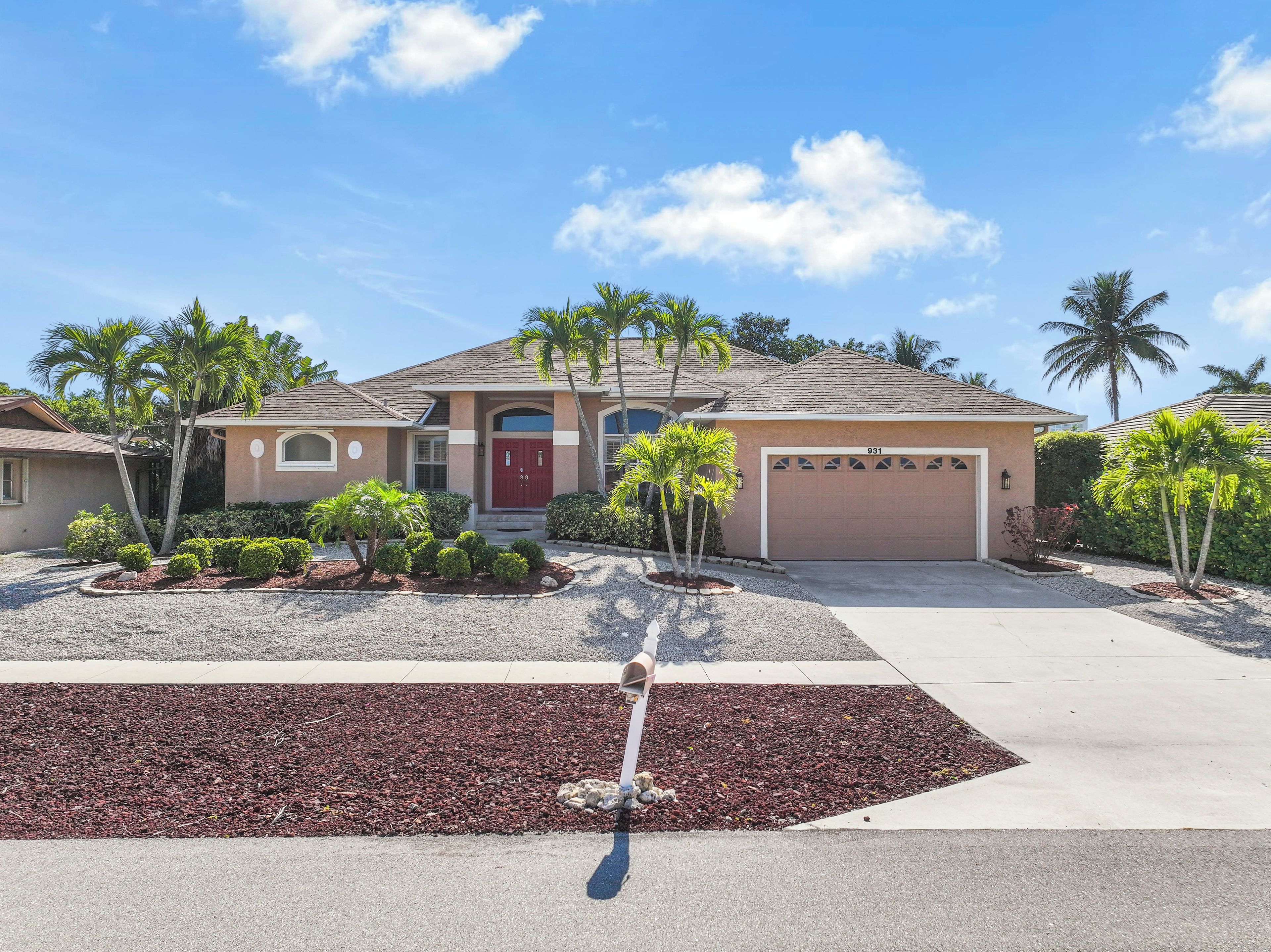 A single-story house with a red door, a garage, and a well-maintained front yard with palm trees and shrubs.