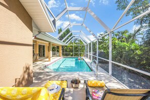 A covered outdoor swimming pool with lounge chairs and a table, surrounded by greenery.
