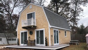 A two-story wooden house with a balcony, sliding glass doors, and a small patio area. - Charming West Harwich Cottage, Newly Renovated, cozy but convenient location (Harwich)
