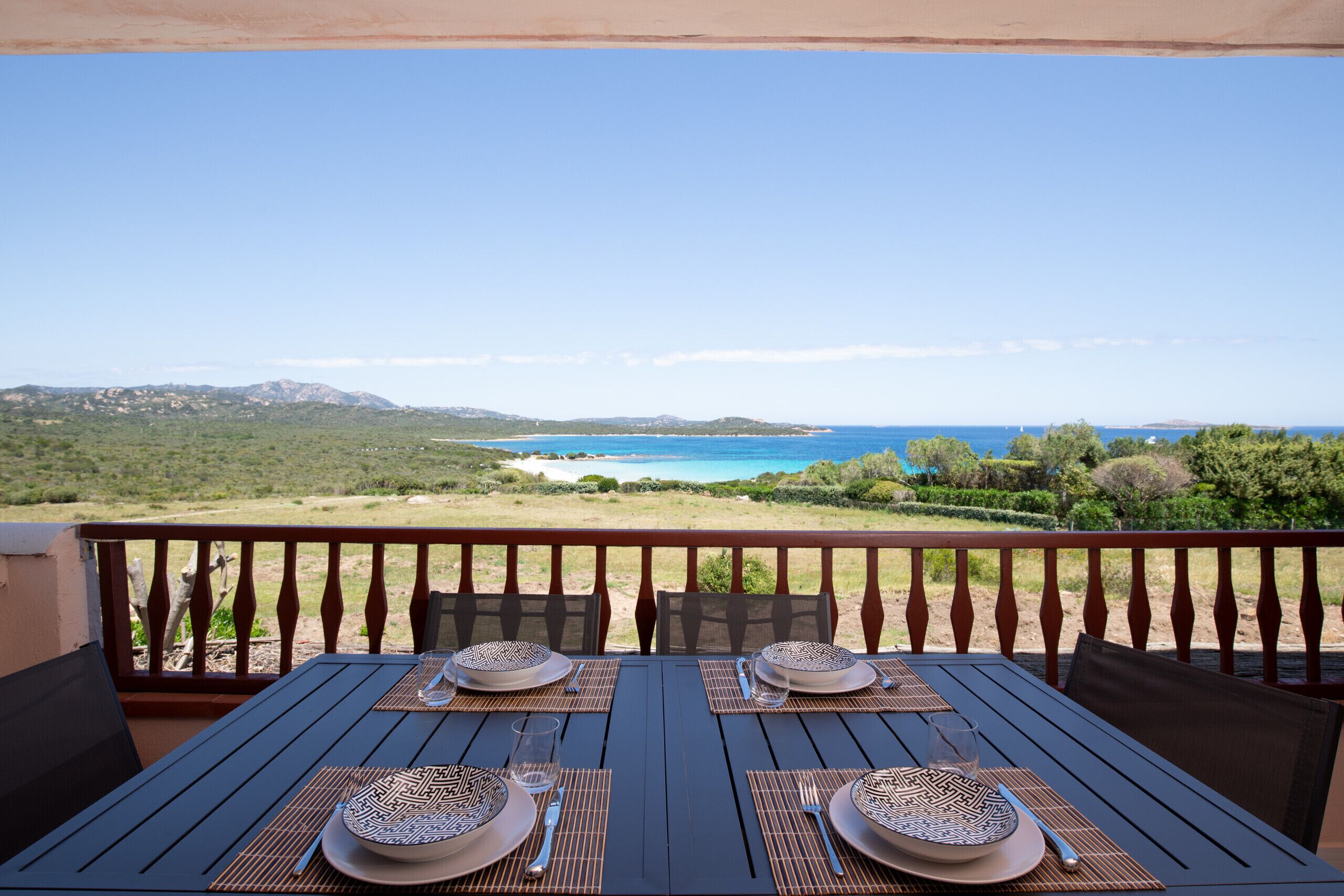 Une table à manger dressée avec des assiettes, des couverts et un verre, sur un balcon donnant sur une plage et des montagnes.