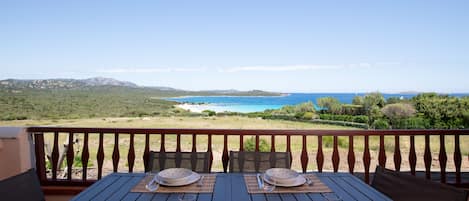 A dining table set with plates, cutlery, and a glass on a balcony overlooking a beach and mountains.